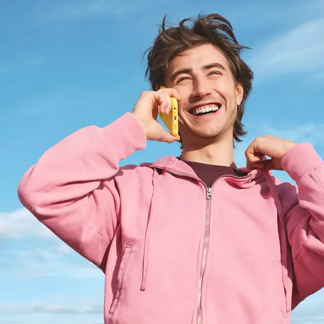 Eine Person steht vor blauem Himmel und telefoniert mit einem gelben Smartphone &ndash; passend zum Stil des neuen Mobilfunkanbieters yelllow.at.