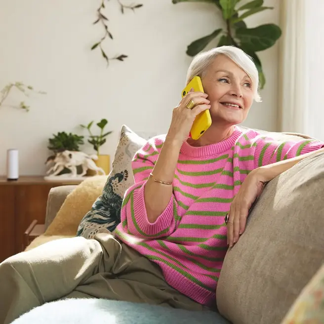 Eine Person sitzt entspannt auf einem Sofa in einem lichtdurchfluteten Wohnzimmer und telefoniert mit einem gelben Smartphone &ndash; ein Moment moderner Kommunikation, wie ihn der Mobilfunkanbieter yelllow.at repr&auml;sentiert.