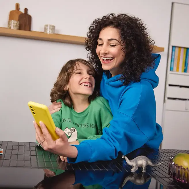 Mutter und ein Kind sitzen in einer K&uuml;che und schauen gemeinsam auf ein gelbes Smartphone &ndash; ein Moment digitaler N&auml;he mit yelllow.at.