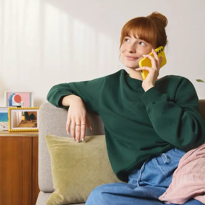 Eine Person sitzt entspannt auf einem Sofa in einem hell eingerichteten Wohnzimmer und telefoniert mit einem gelben Smartphone &ndash; ein Bild f&uuml;r einfache Kommunikation mit yelllow.at.